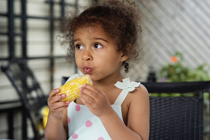 Mixed-race toddler eating corn-on-the-cob outdoors.